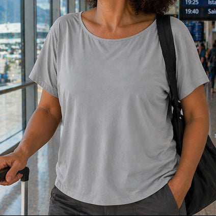 Woman with a suitcase in an airport terminal, looking out of a large window wearing the eclipse boatneck top in pewter.