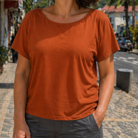 Woman in a sienns eclipse boatneck shirt and straw hat standing on a street with shops and mountains in the background