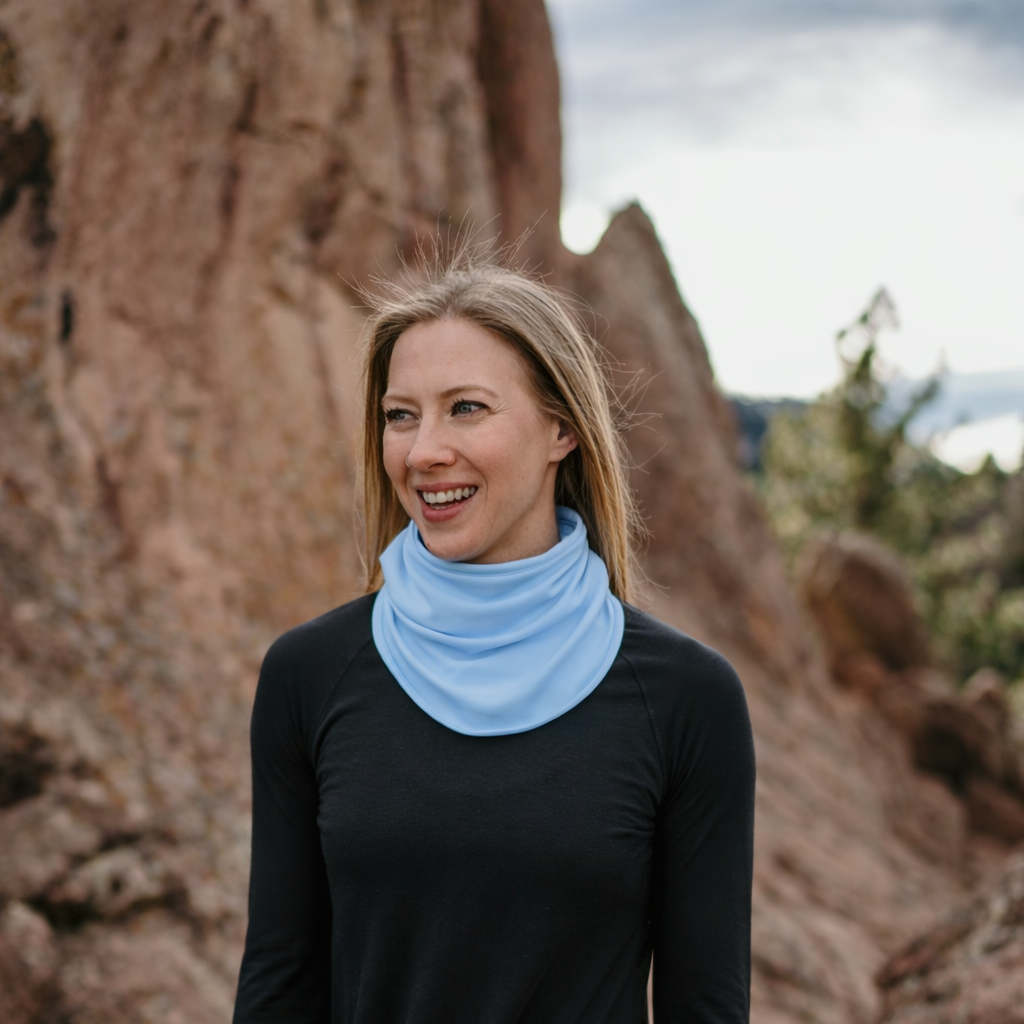 Woman wearing a light blue eclipse neck gaiter standing in front of rocky terrain.